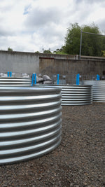 Row of currogated metal planters on a gravel surface with a cloudy sky in the background.