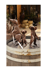 Three metal meerkat sculptures on a rustic wooden barrel with decorative elements in the background.