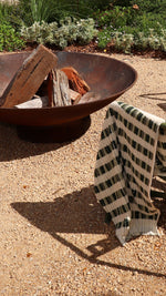 Rustic metal fire pit with wood and a checkered blanket hanging on the arm of a garden chair on a gravel surface.