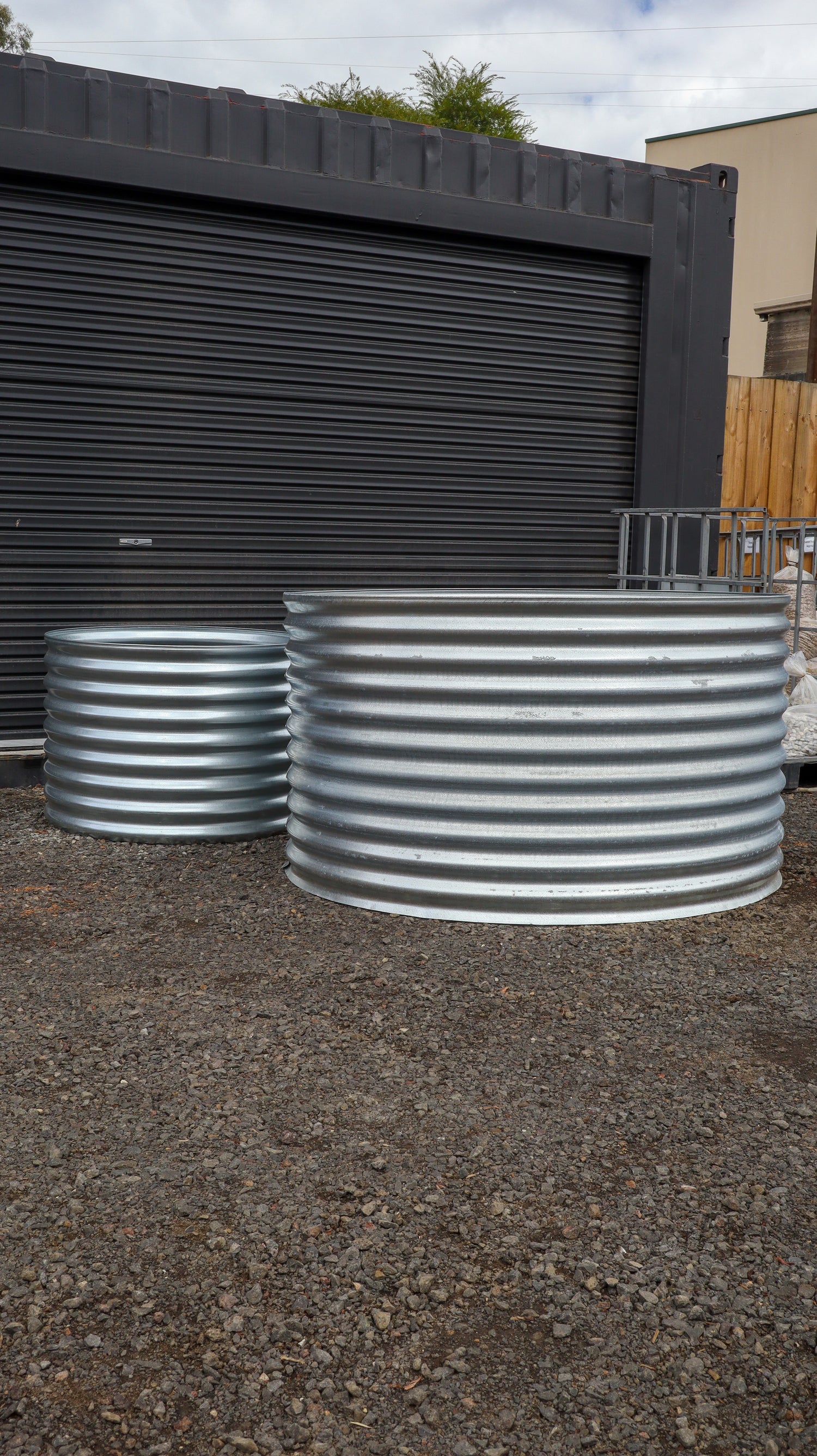 Two large metal water tanks on a gravel surface with a storage unit in the background.