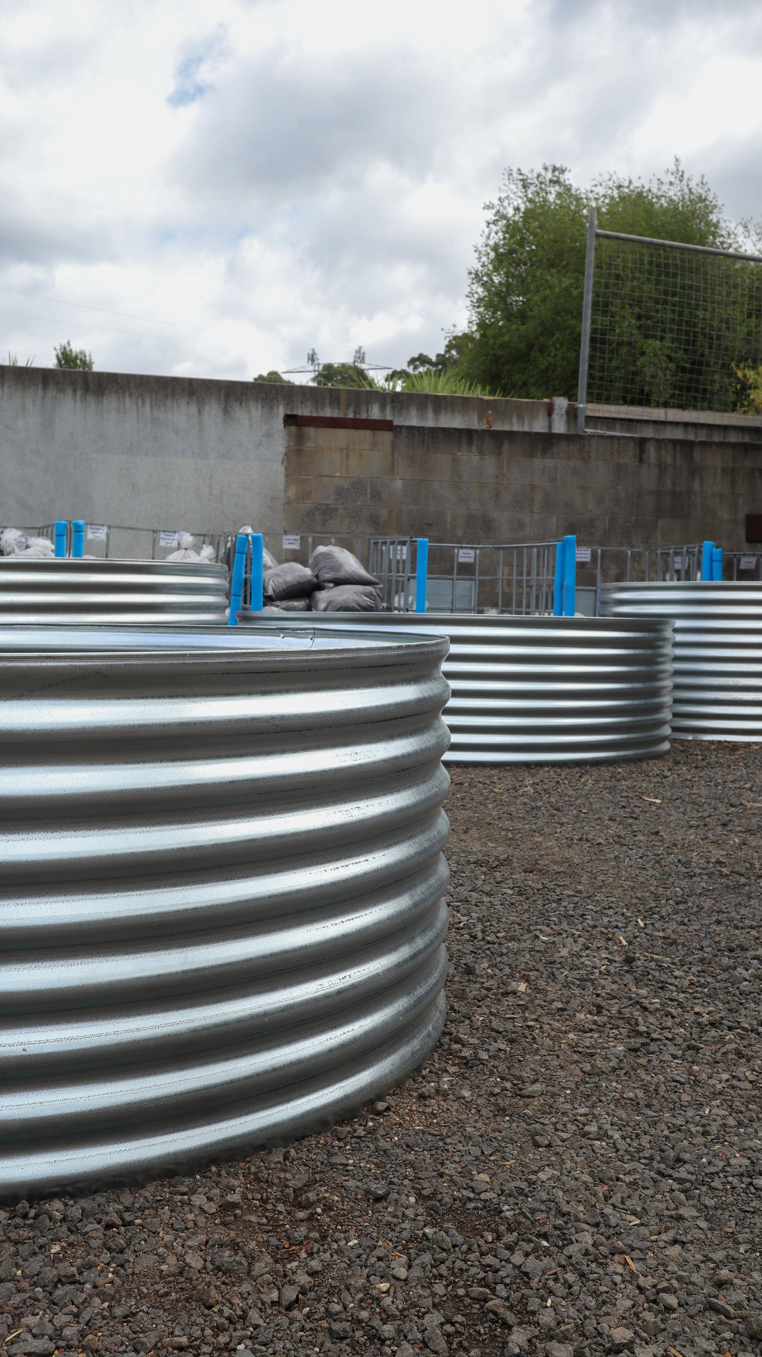 Row of currogated metal planters on a gravel surface with a cloudy sky in the background.