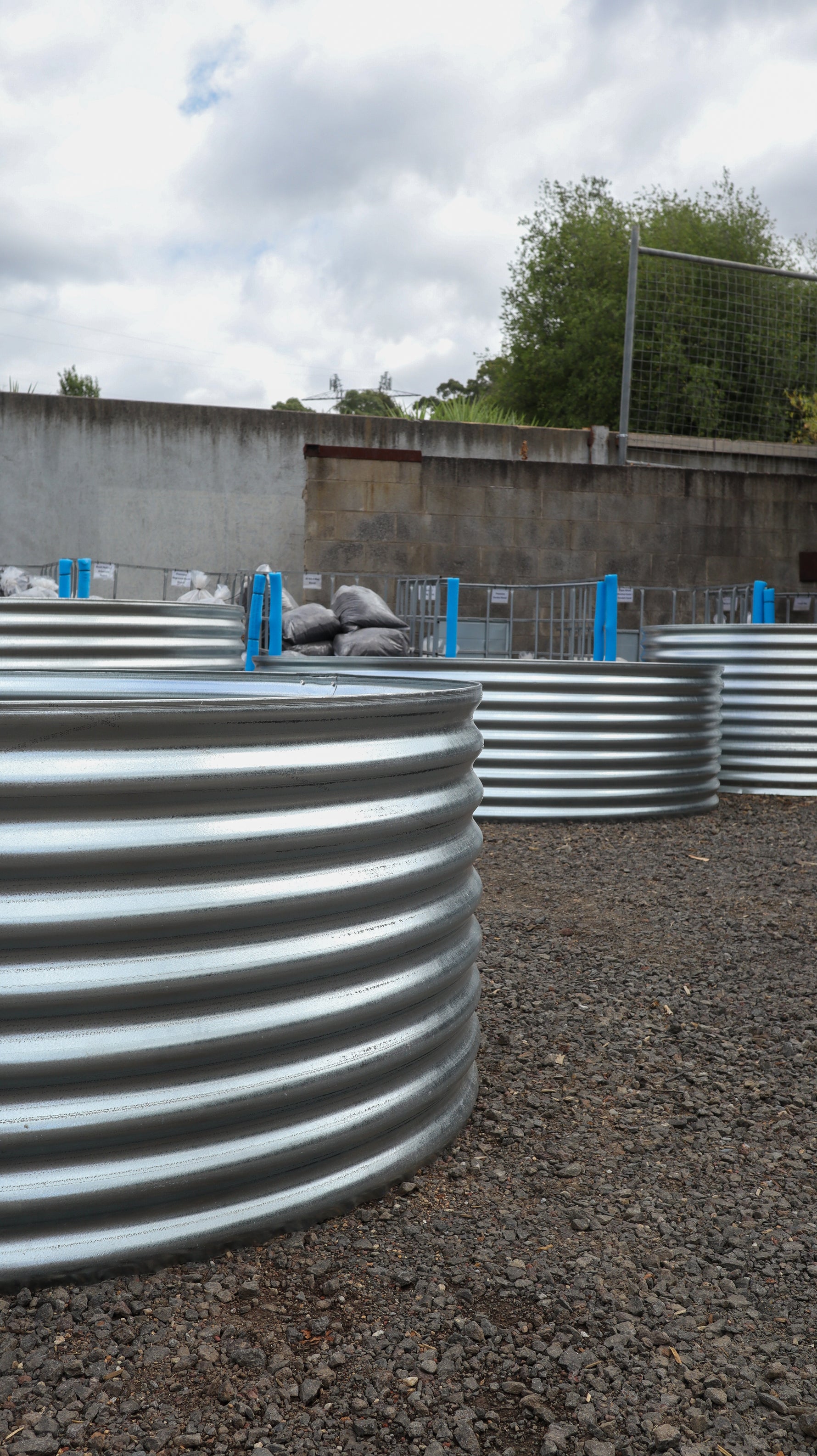 Row of currogated metal planters on a gravel surface with a cloudy sky in the background.