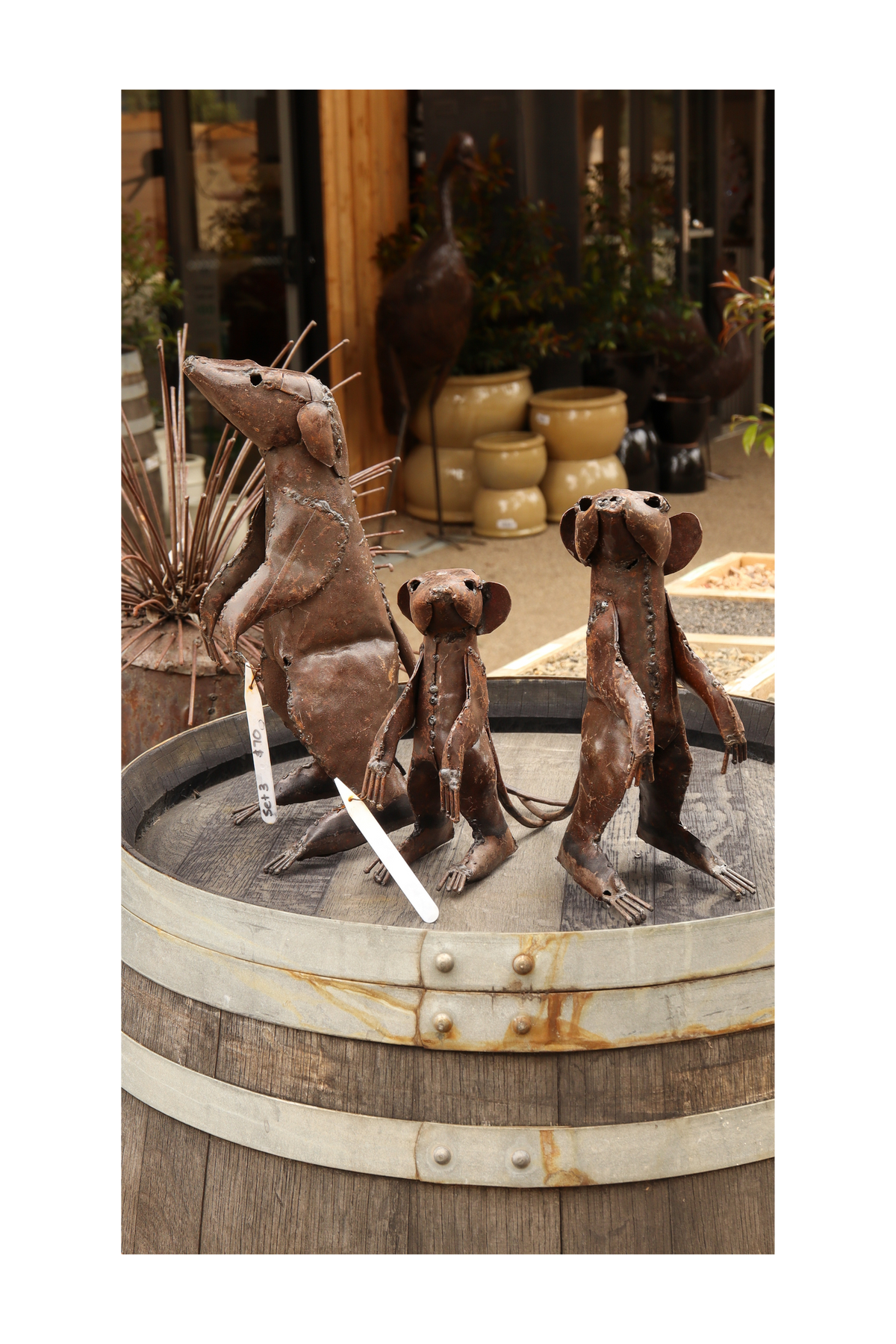 Three metal meerkat sculptures on a rustic wooden barrel with decorative elements in the background.